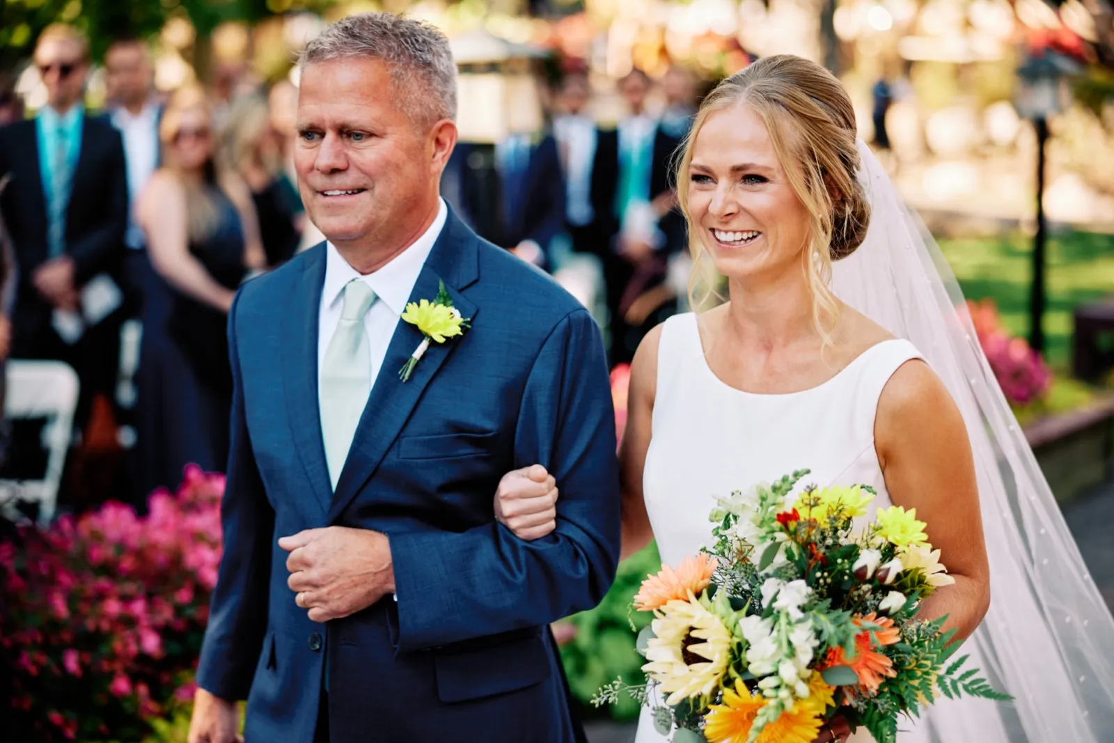 The bride smiles broadly while walking down the outdoor ceremony aisle on her father's arm, holding a vibrant sunflower and orange gerbera bouquet. — Tim Larsen Photography, Brainerd Lakes MN