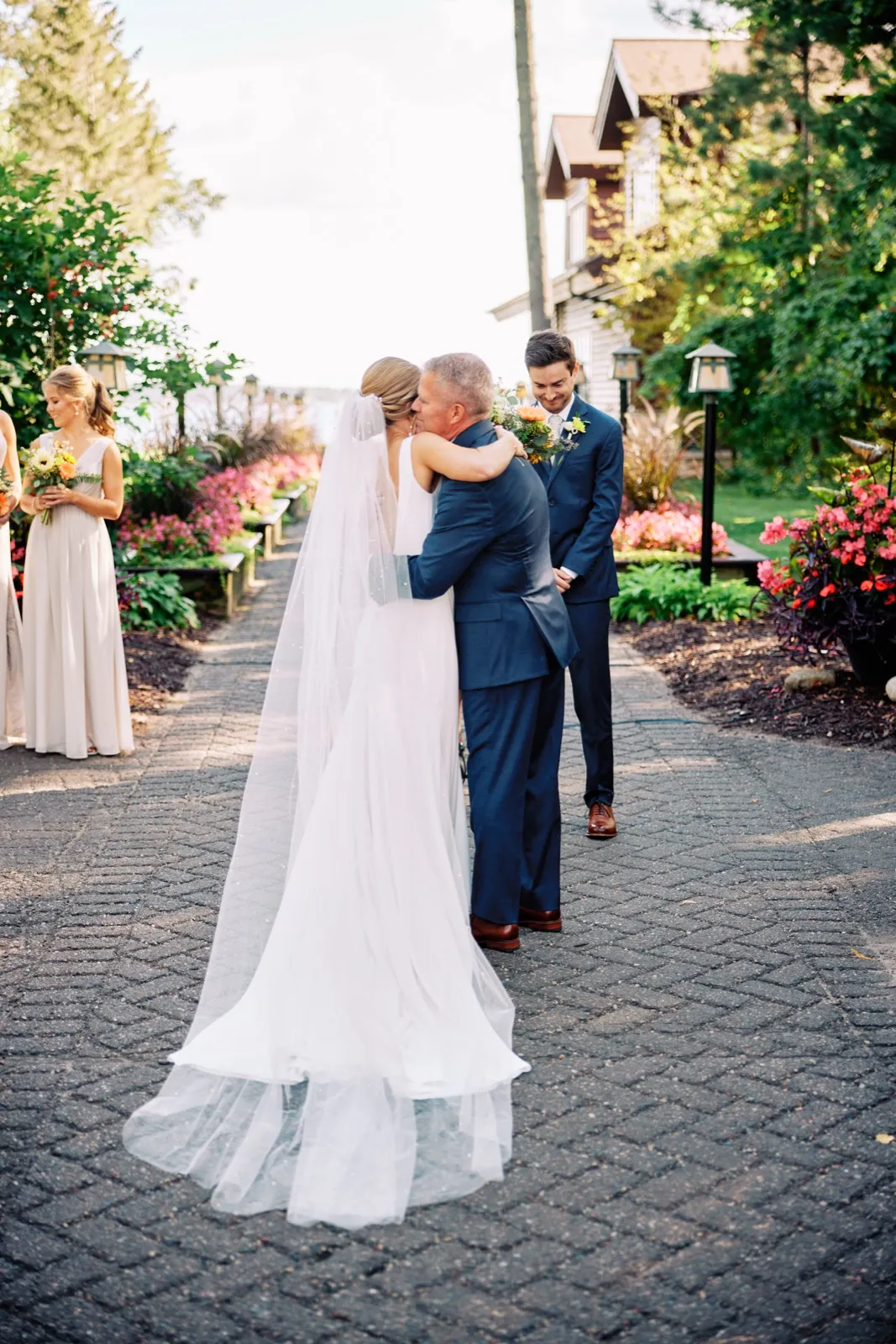 The bride embraces her father at the end of the aisle as the groom waits and watches, her long veil trailing across a brick path lined with flowers at Grand View Lodge. — Tim Larsen Photography, Brainerd Lakes MN