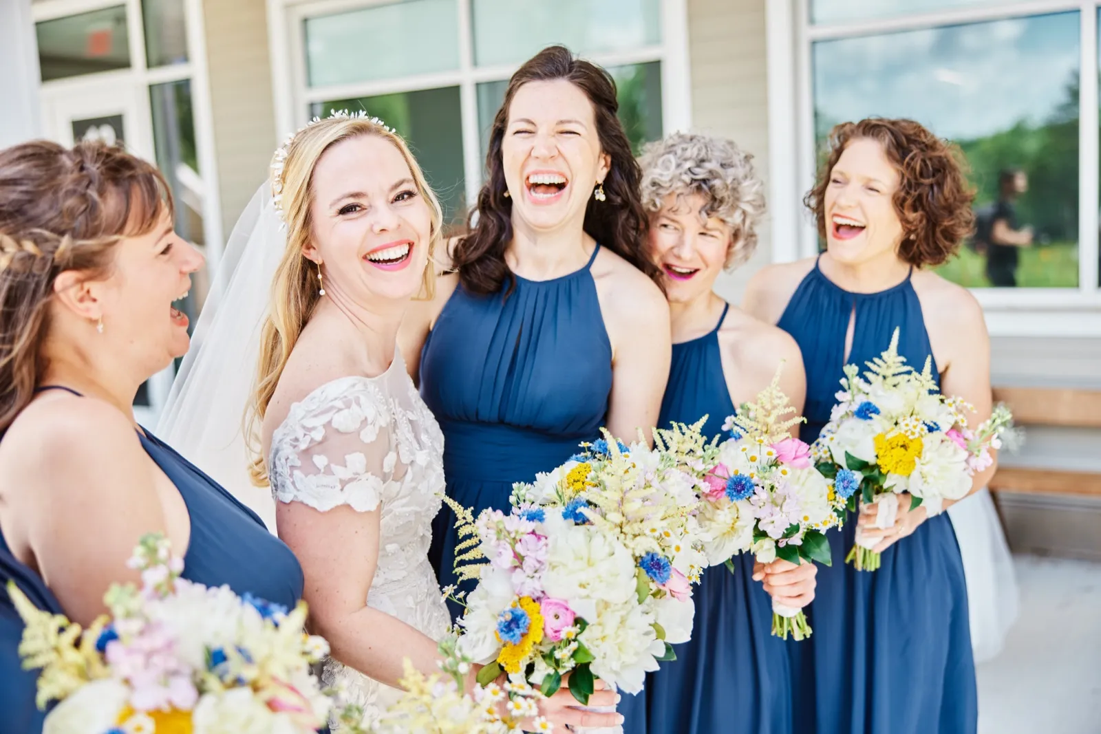 The bride and her bridesmaids in dusty blue dresses laugh together while holding wildflower bouquets outside the venue. — Tim Larsen Photography, Minnesota