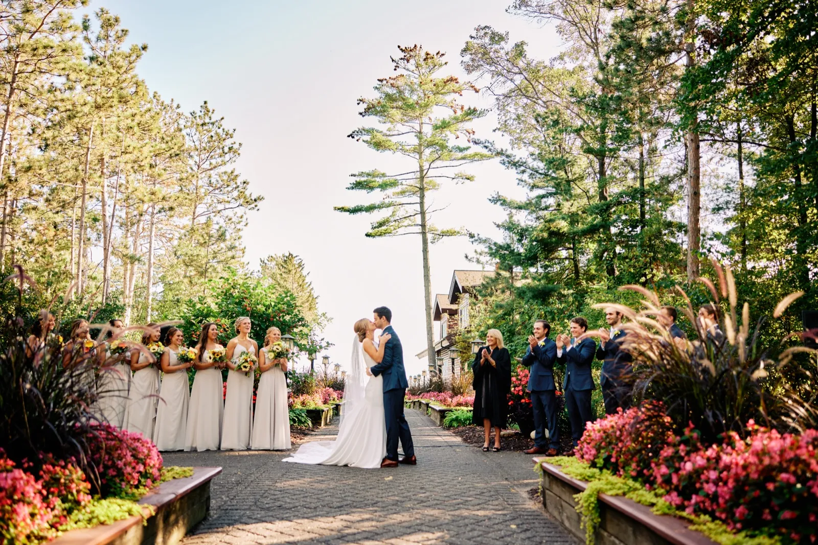 The bride and groom share their first kiss at the end of an outdoor garden aisle while the bridal party and officiant applaud beneath towering pine trees at Grand View Lodge. — Tim Larsen Photography, Brainerd Lakes MN