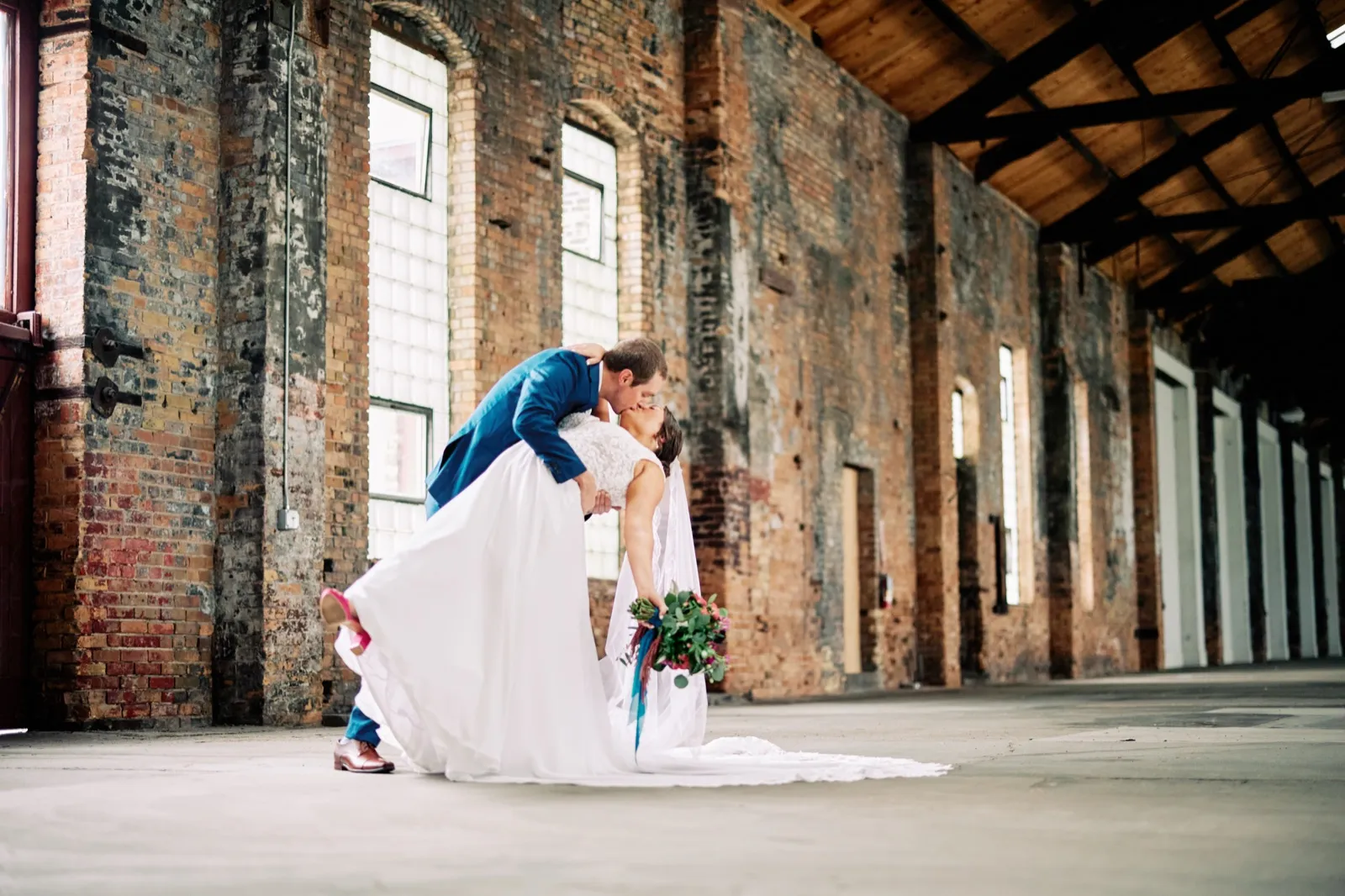 Craig dips Maggie into a kiss beside tall brick columns and industrial windows, her lace train and bouquet ribbons sweeping toward the floor — Tim Larsen Photography, Brainerd Lakes MN
