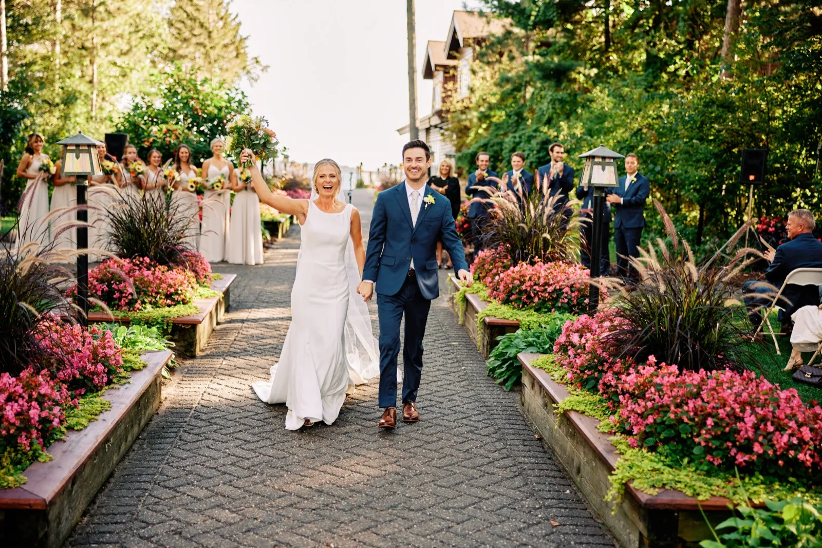The bride raises her bouquet overhead and laughs as she walks hand-in-hand with the groom down a brick garden aisle lined with pink flowers and the bridal party. — Tim Larsen Photography, Brainerd Lakes MN