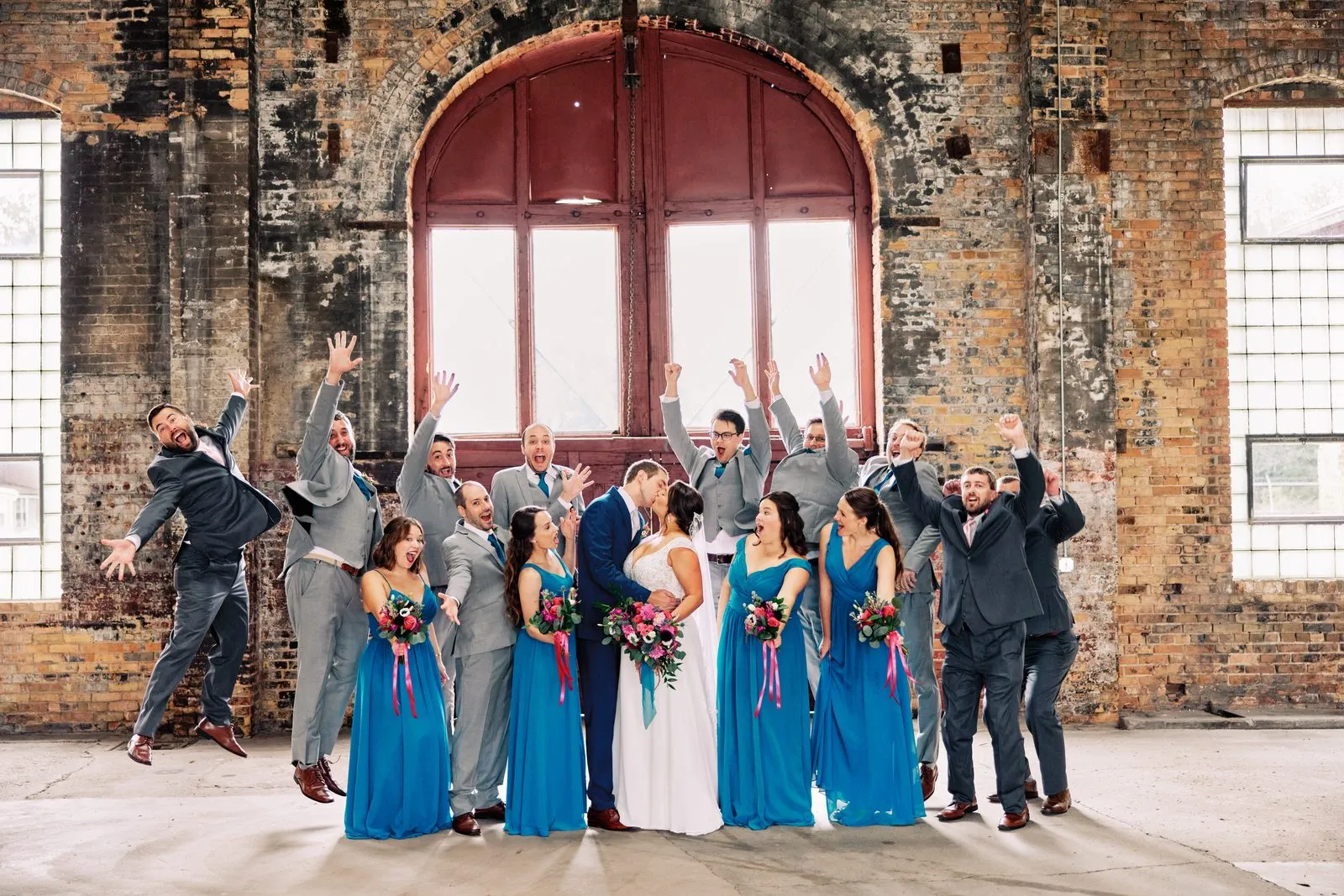 Maggie and Craig kiss beneath an arched red freight door while their wedding party jumps and cheers around them — Tim Larsen Photography, Brainerd Lakes MN