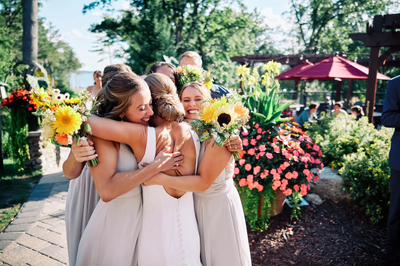 The bride and her bridesmaids share a tight group hug holding sunflower bouquets on a sunlit garden patio with red umbrellas and bright florals behind them. — Tim Larsen Photography, Brainerd Lakes MN