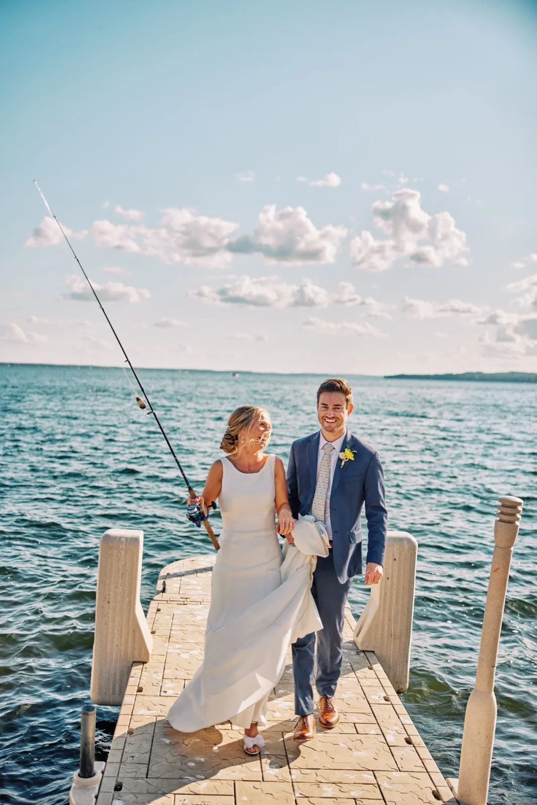 The bride holds a fishing rod aloft as she and the groom walk together down a dock with Gull Lake stretching behind them. — Tim Larsen Photography, Brainerd Lakes MN