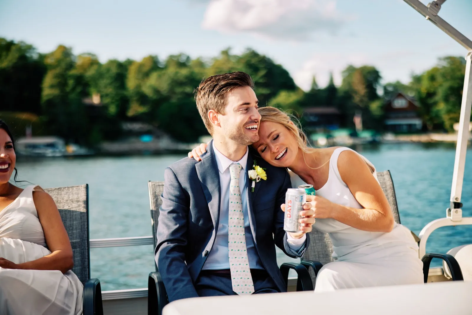 The bride leans her head against the groom's shoulder, both laughing, while seated on a pontoon boat on a sunlit lake with a bridesmaid laughing beside them. — Tim Larsen Photography, Brainerd Lakes MN