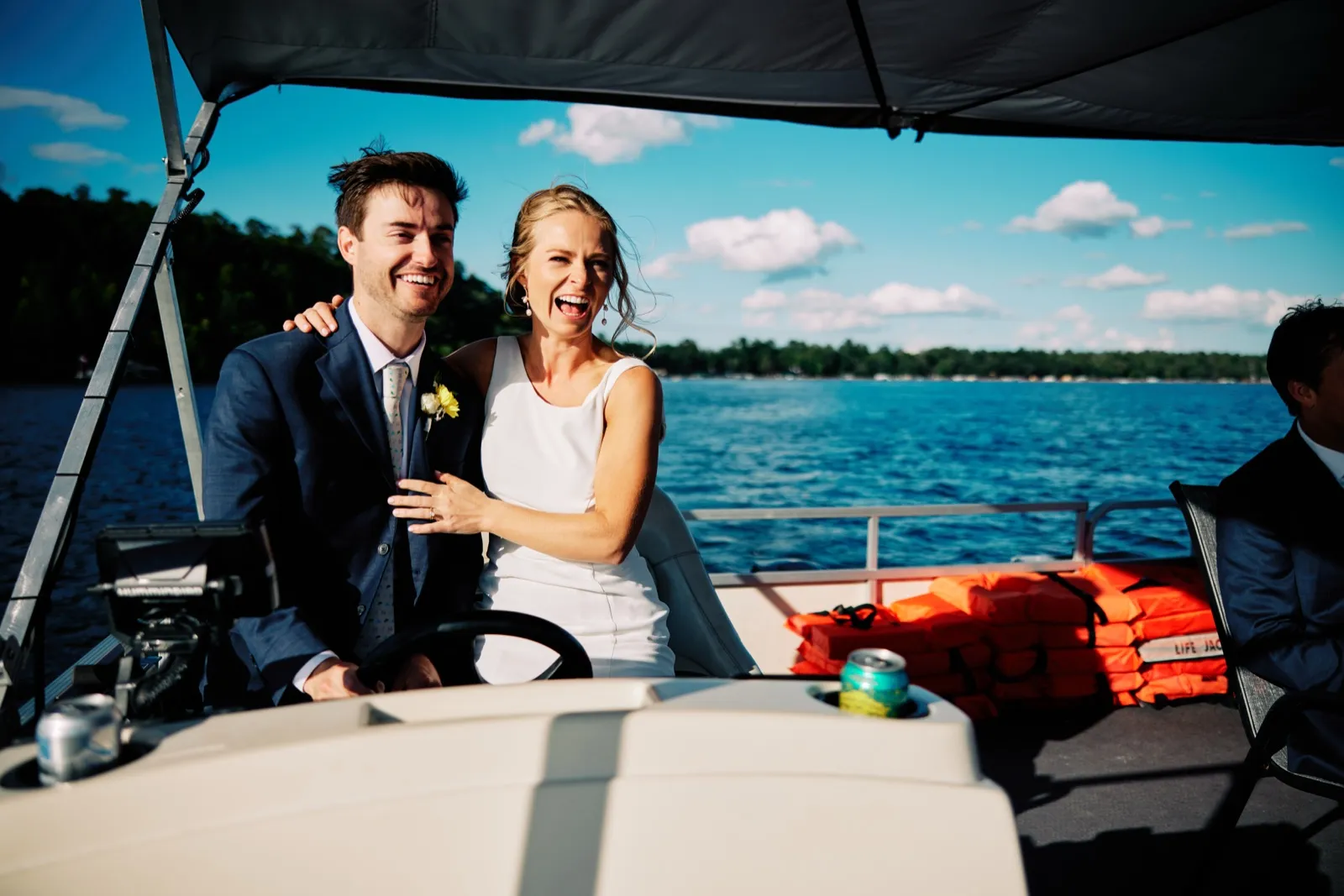 The bride and groom laugh together at the wheel of a pontoon boat on a sunlit lake, with stacked orange life jackets and another guest visible to the side. — Tim Larsen Photography, Brainerd Lakes MN