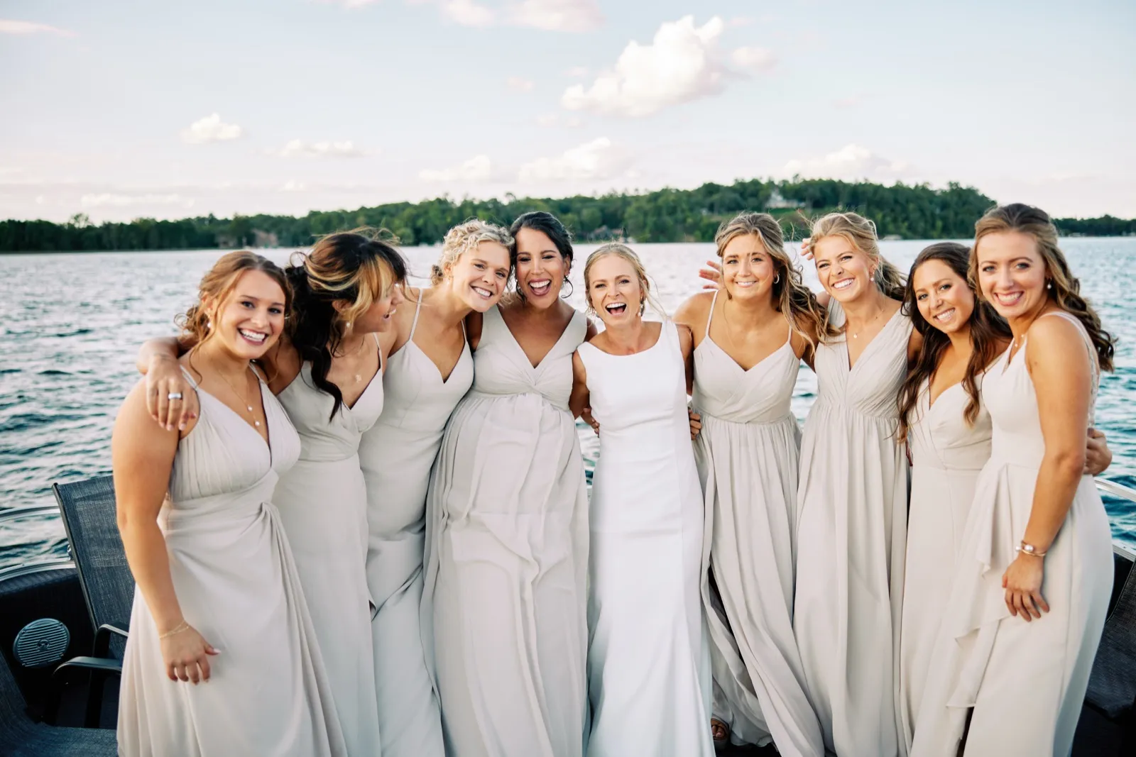 The bride and eight bridesmaids in pale neutral gowns laugh together on a pontoon boat with Gull Lake and a tree-lined shore behind them. — Tim Larsen Photography, Brainerd Lakes MN