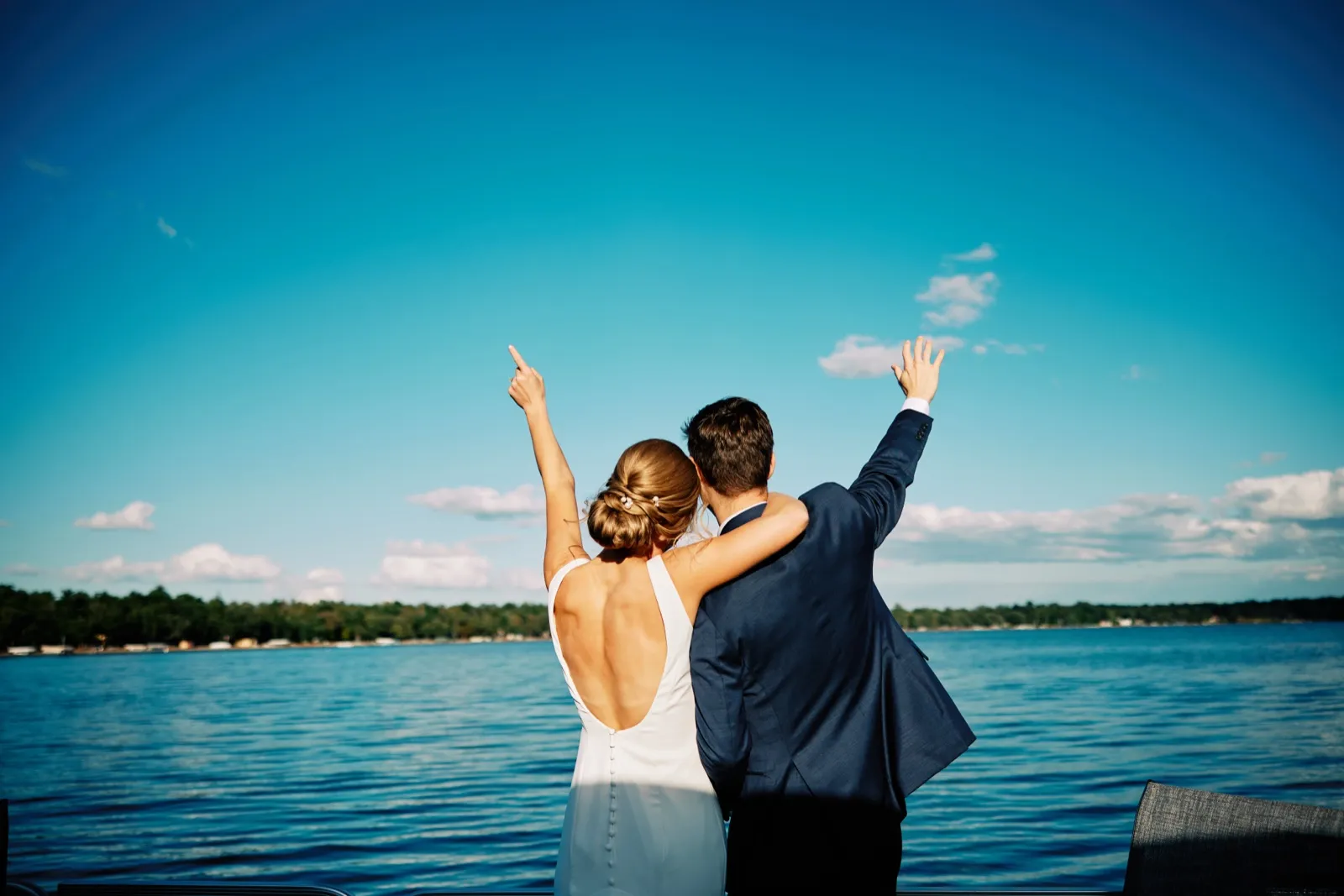 The bride and groom stand together facing Gull Lake with their arms raised in celebration against a vast blue sky. — Tim Larsen Photography, Brainerd Lakes MN