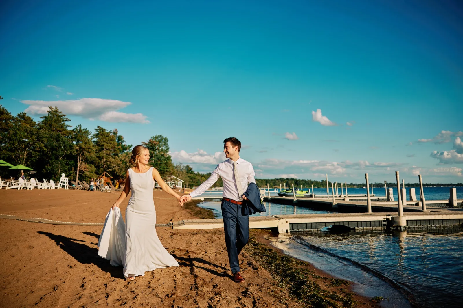 The bride lifts her gown as she walks hand-in-hand with the groom along a sandy lakeshore, with Grand View Lodge's docks extending into Gull Lake behind them. — Tim Larsen Photography, Brainerd Lakes MN