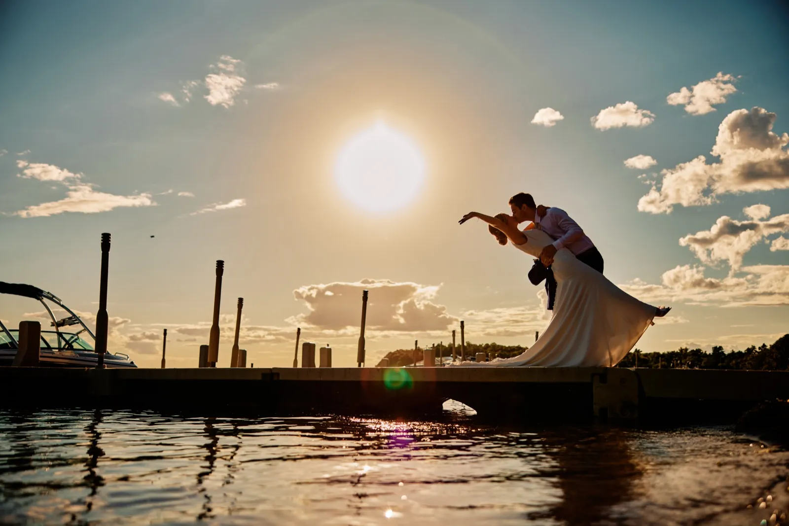 The groom dips the bride into a kiss at the end of a dock, silhouetted against a low sun over the lake with boats and pylons lining the marina. — Tim Larsen Photography, Brainerd Lakes MN
