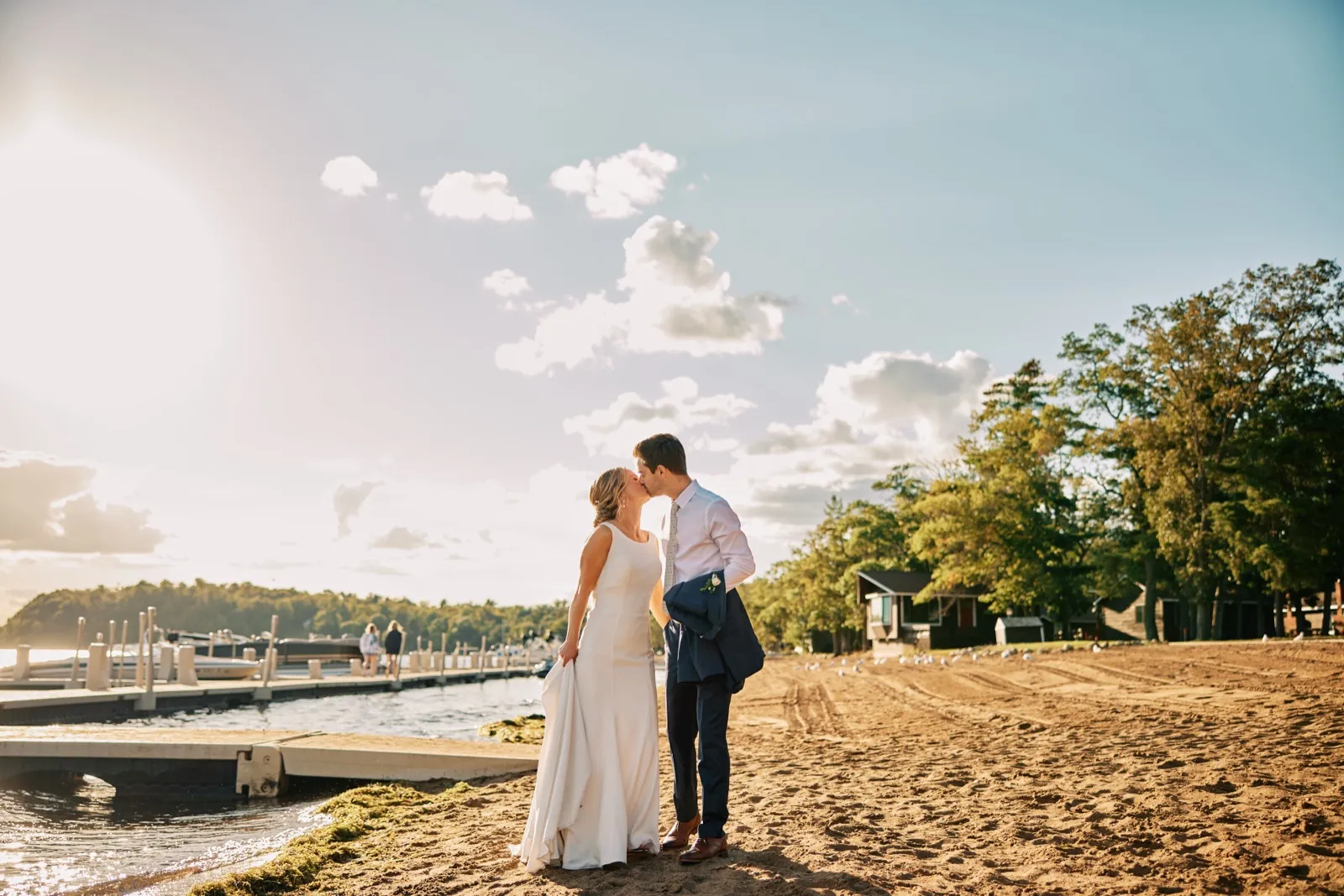The bride and groom kiss on the sandy beach at Grand View Lodge with the dock, lake, and lodge cabins visible behind them in golden evening light. — Tim Larsen Photography, Brainerd Lakes MN