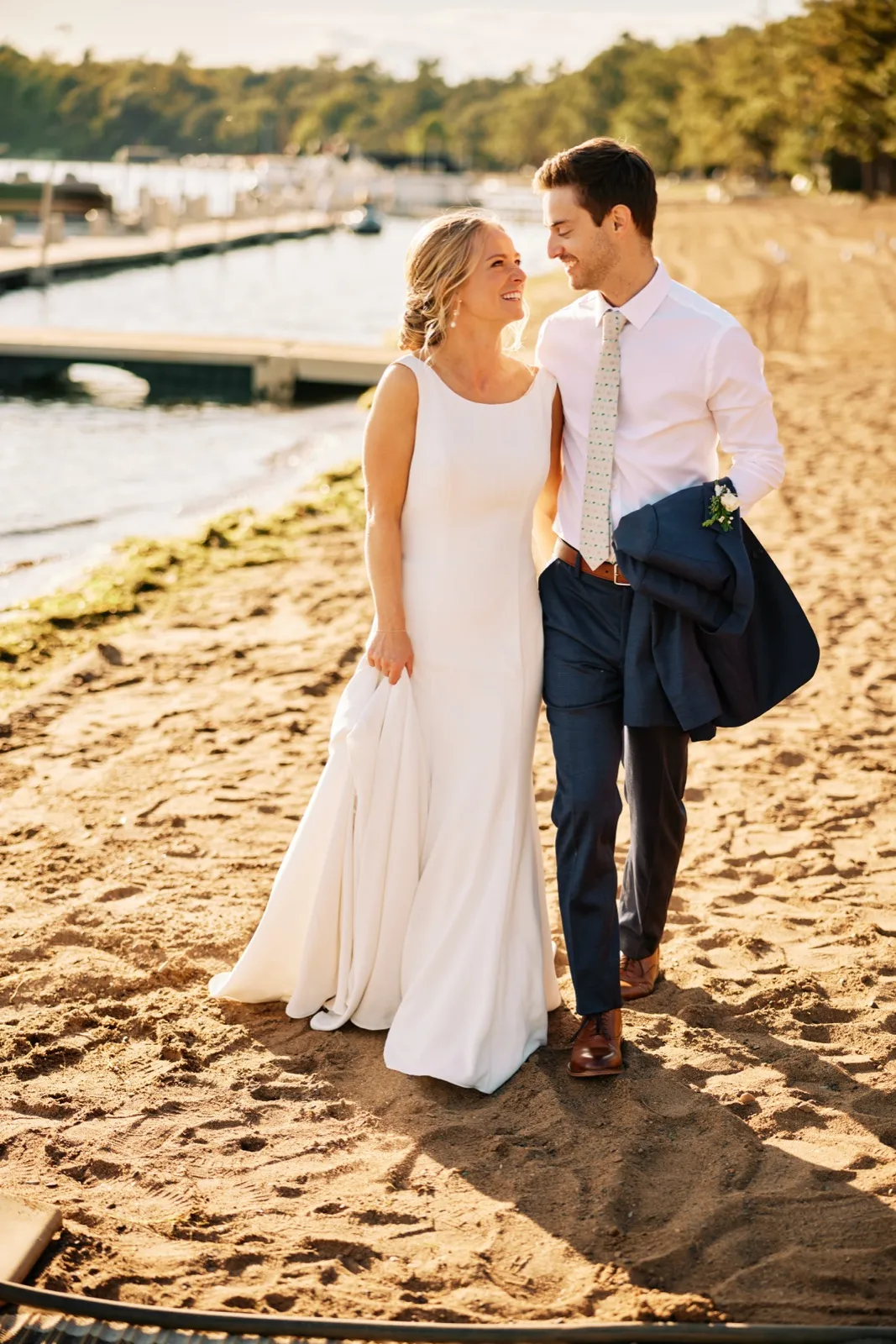 The bride and groom walk together on a sandy beach in golden evening light, with a marina and Gull Lake softly visible behind them. — Tim Larsen Photography, Brainerd Lakes MN