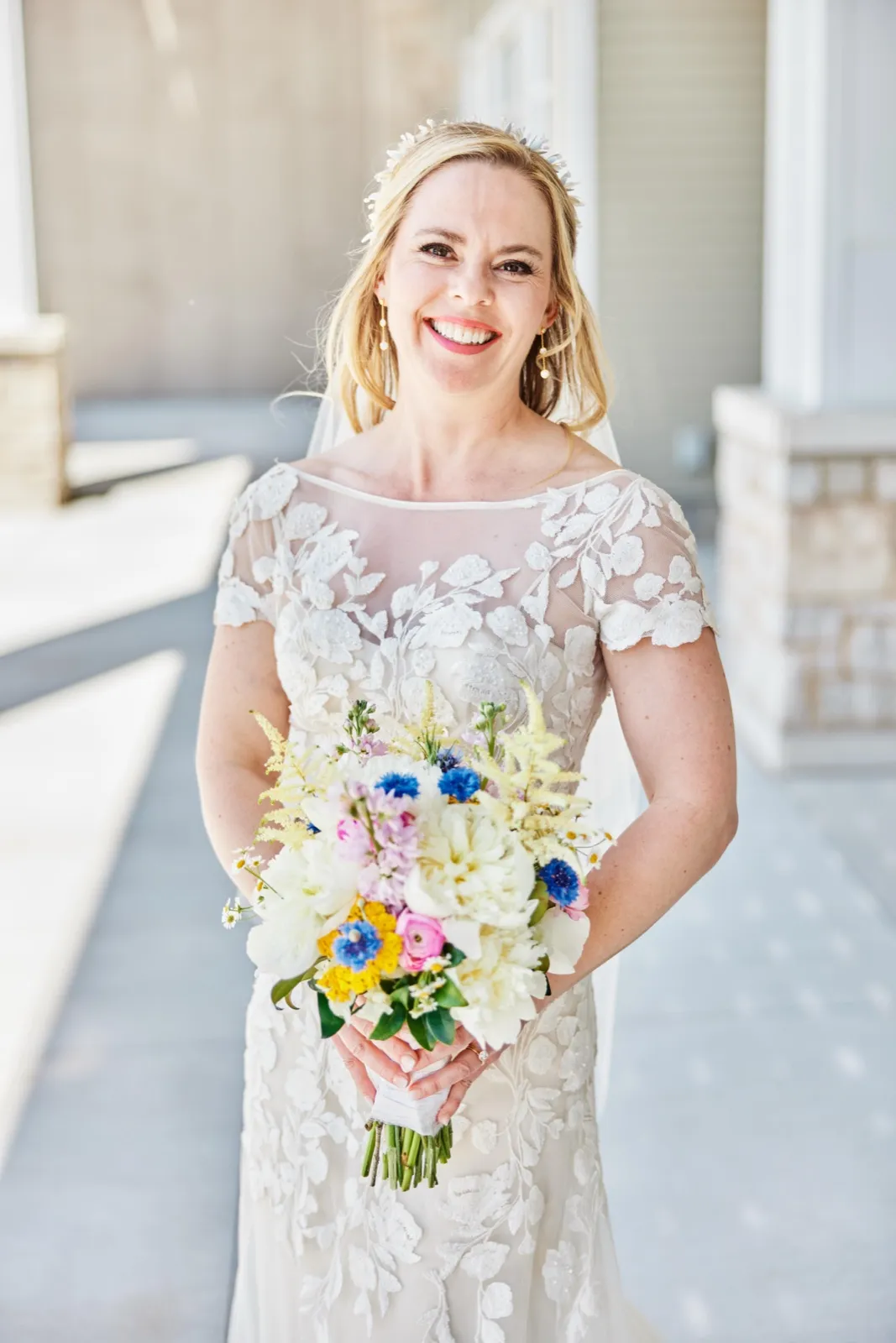 The bride smiles broadly at the camera while holding a colorful wildflower bouquet, wearing a lace gown with sheer floral bodice on a shaded stone porch. — Tim Larsen Photography, Minnesota