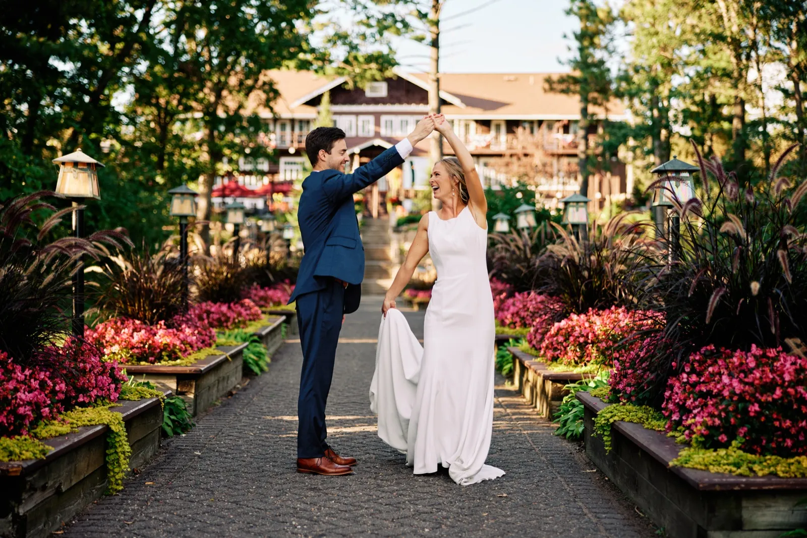 The groom twirls the bride on a symmetrical garden path lined with pink flowers and lanterns, with the Grand View Lodge main building rising behind them in evening light. — Tim Larsen Photography, Brainerd Lakes MN