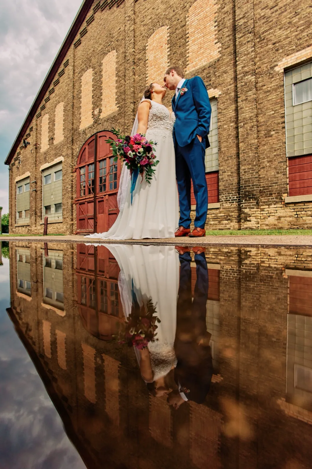 Maggie and Craig kiss outside a historic brick industrial building, their figures mirrored in a puddle on the pavement below — Tim Larsen Photography, Brainerd Lakes MN
