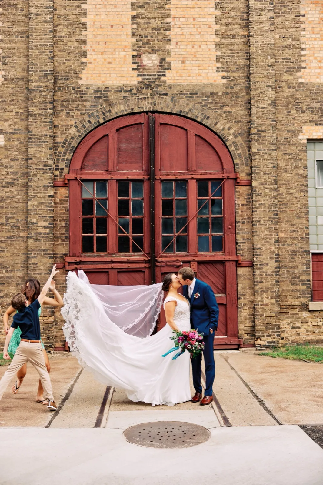 Maggie and Craig kiss in front of the towering arched red doors of Northern Pacific Center while helpers hold her long veil out to the wind — Tim Larsen Photography, Brainerd Lakes MN