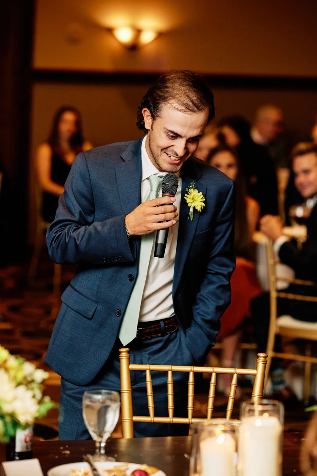 The best man laughs and looks down while holding a microphone during a toast, with seated guests reacting in the softly lit reception background.