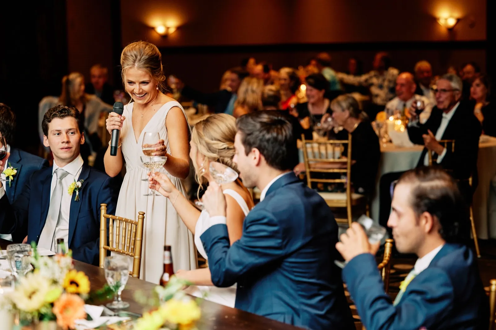 The bride holds a microphone and a wine glass while addressing guests at her reception, with the groom and bridal party reacting at the head table. — Tim Larsen Photography, Brainerd Lakes MN