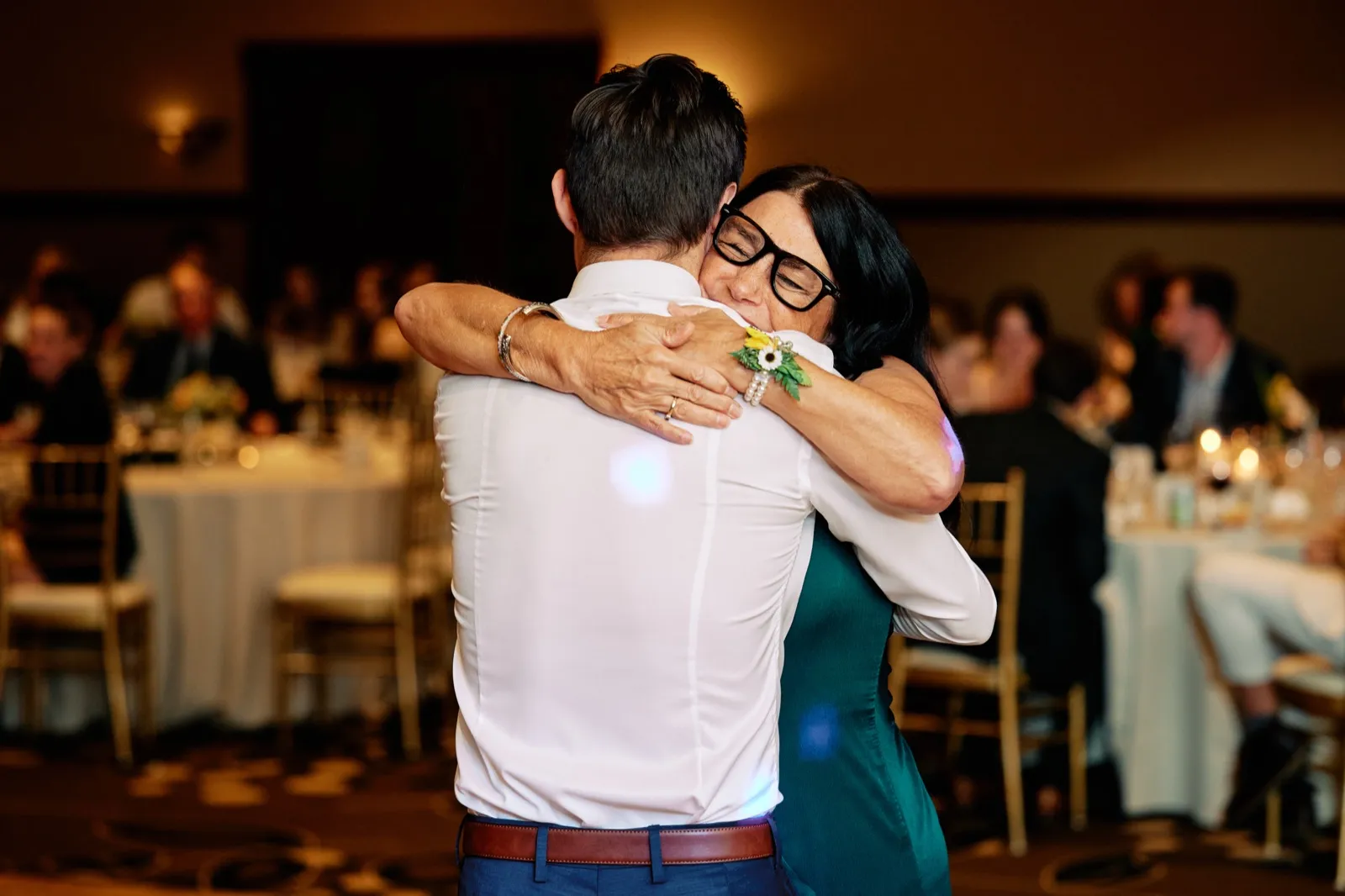 The groom's mother closes her eyes and holds him tightly during their dance, her sunflower corsage visible on his back as guests watch from tables behind them. — Tim Larsen Photography, Brainerd Lakes MN