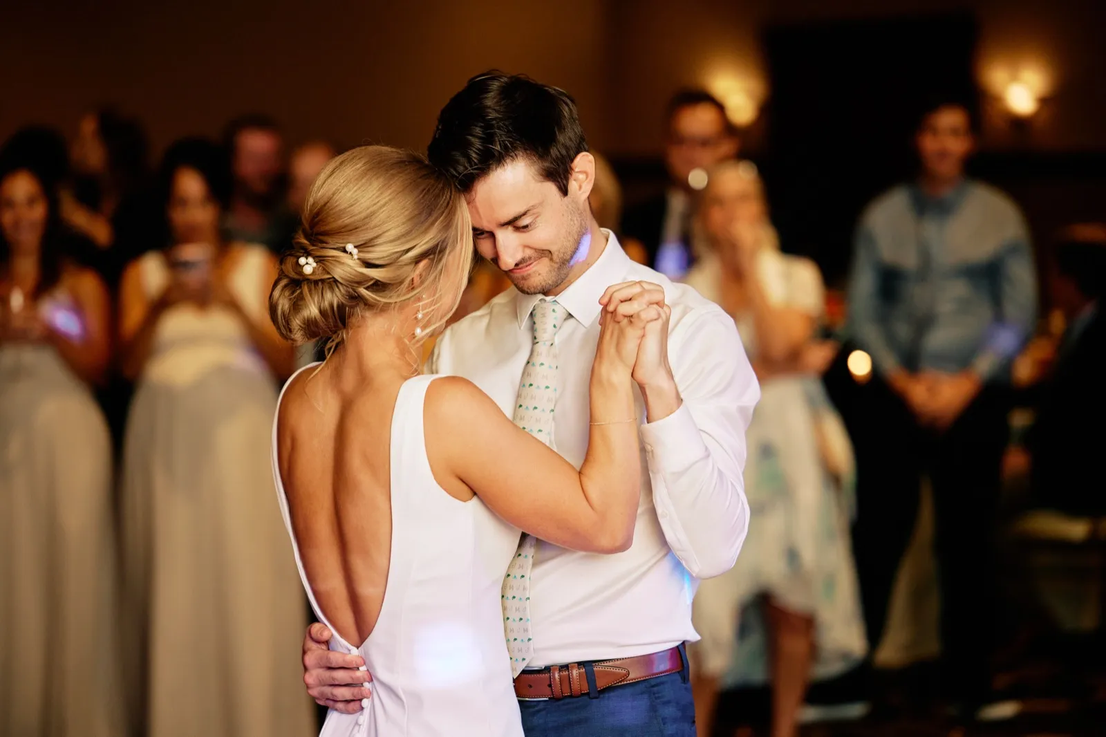 The groom rests his forehead against the bride's temple as they hold clasped hands during their first dance, guests in soft focus behind them. — Tim Larsen Photography, Brainerd Lakes MN