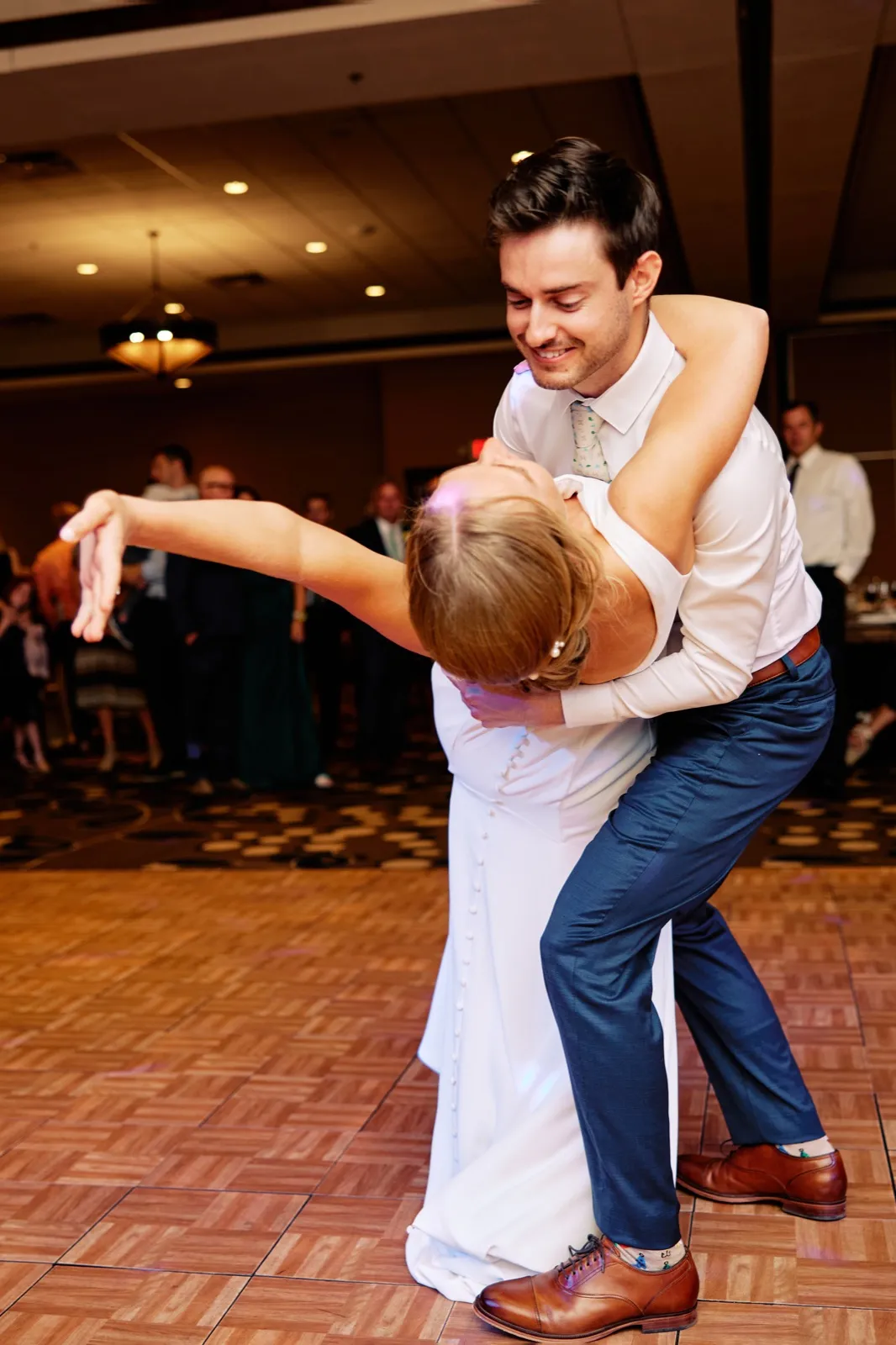 The groom dips the bride backward on the parquet ballroom dance floor while bridesmaids and guests watch from the edges