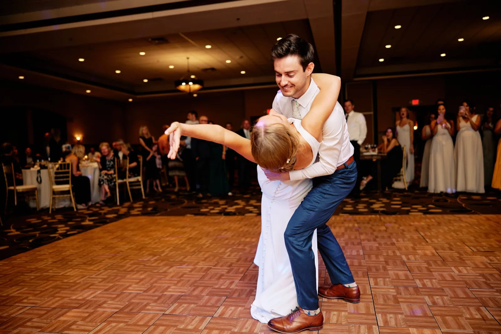 The groom dips the bride backward on the parquet Grand Ballroom dance floor while bridesmaids and guests watch from the edges. — Tim Larsen Photography, Brainerd Lakes MN