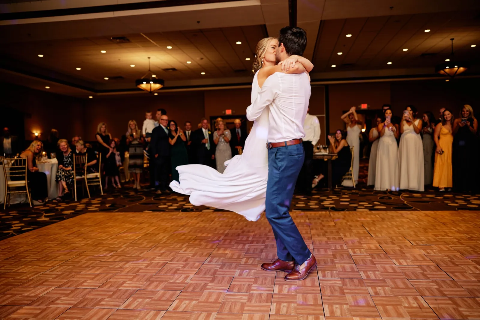 The groom lifts and spins the bride during their first dance as guests and the bridal party watch from around the ballroom floor. — Tim Larsen Photography, Brainerd Lakes MN