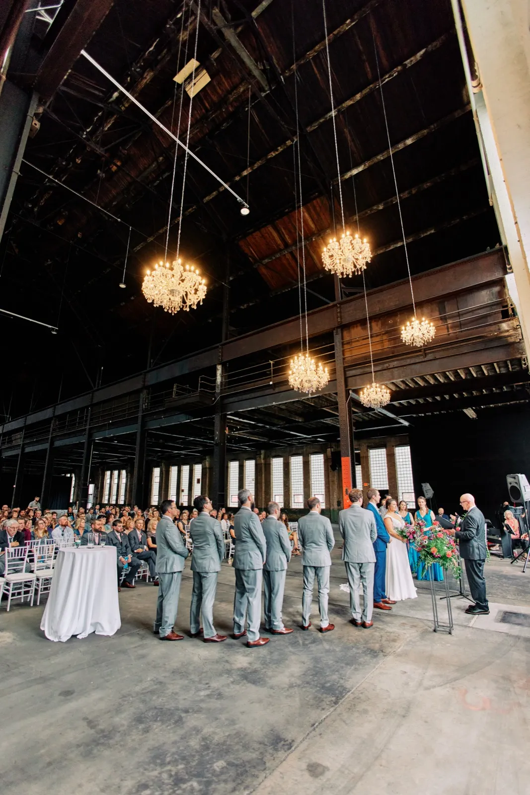 A wide view from behind the wedding party shows Maggie and Craig's ceremony beneath chandeliers and the hall's soaring exposed trusses — Tim Larsen Photography, Brainerd Lakes MN