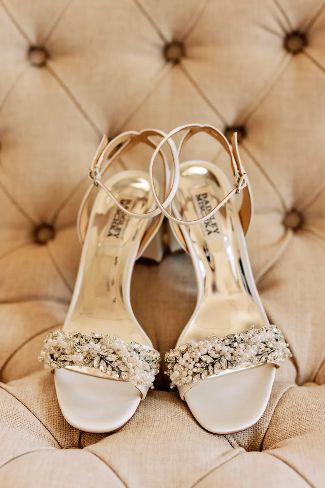 A pair of pearl- and crystal-embellished Badgley Mischka bridal heels staged symmetrically on a tufted champagne-colored cushion. — Tim Larsen Photography, Twin Cities MN