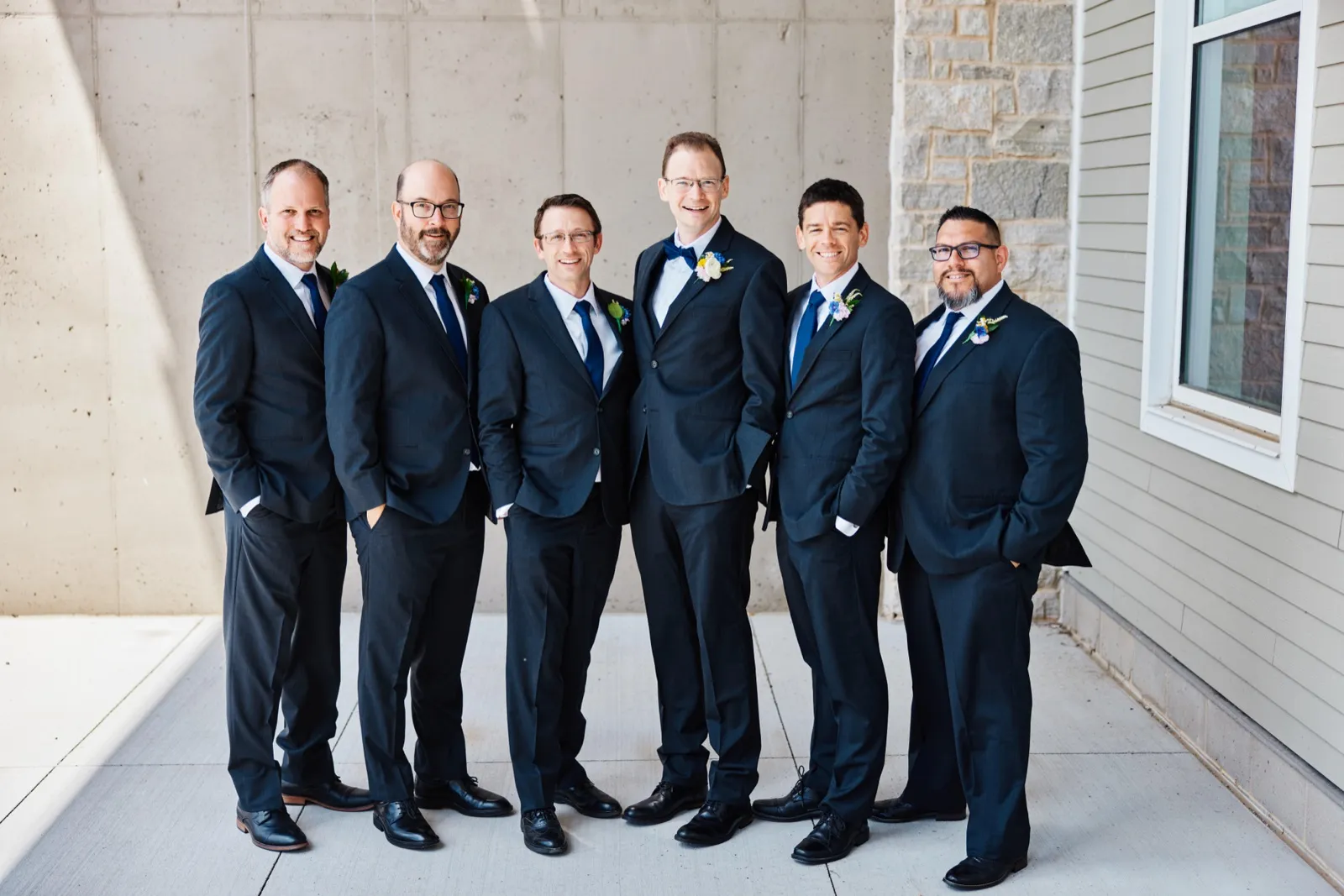 The groom stands center with his five groomsmen in matching navy suits, smiling at the camera against a concrete and stone wall. — Tim Larsen Photography, Minnesota