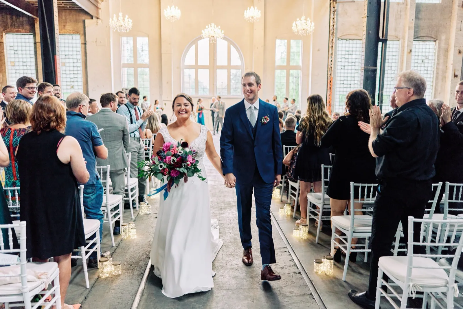 Maggie and Craig hold hands and walk down the aisle while guests in chiavari chairs applaud beneath chandeliers and tall arched windows — Tim Larsen Photography, Brainerd Lakes MN