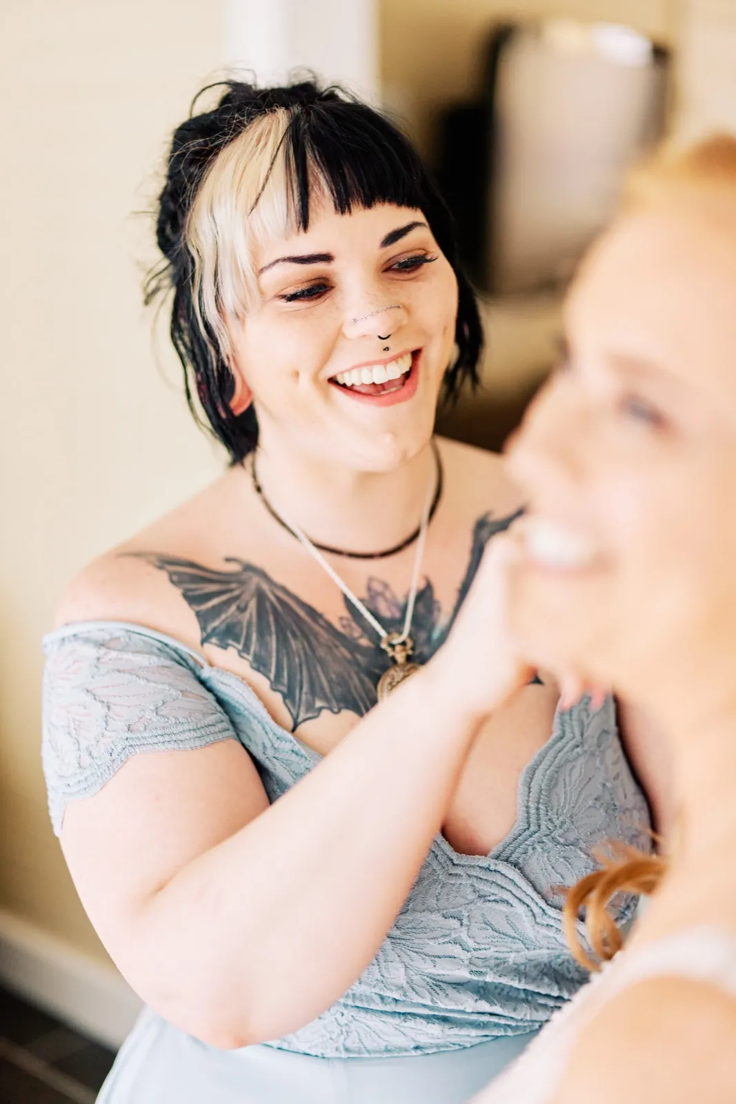A bridesmaid with two-tone hair and a chest tattoo laughs openly while sharing a moment with the bride, who is visible out of focus in the foreground. — Tim Larsen Photography, Brainerd Lakes MN