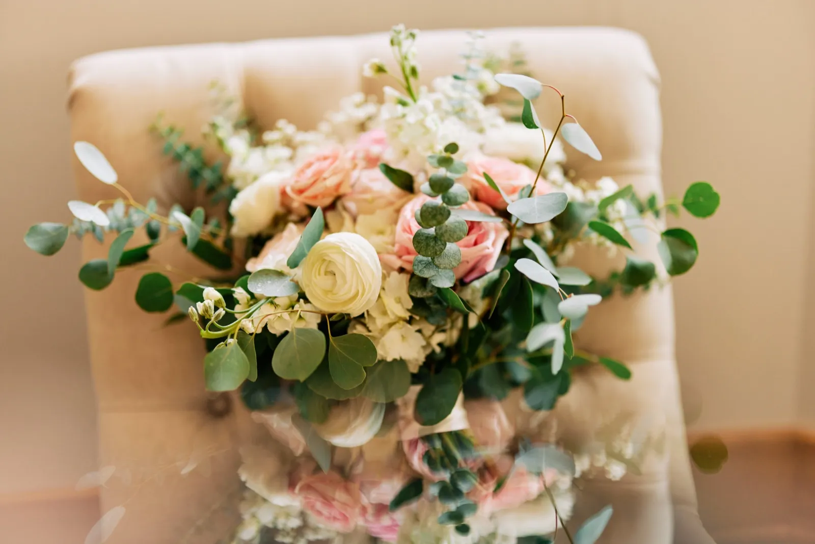 A bridal bouquet of white ranunculus, blush garden roses, and trailing eucalyptus rests on a tufted cream chair in soft window light, with a faint double-exposure overlay of the bouquet beneath. — Tim Larsen Photography, Twin Cities MN