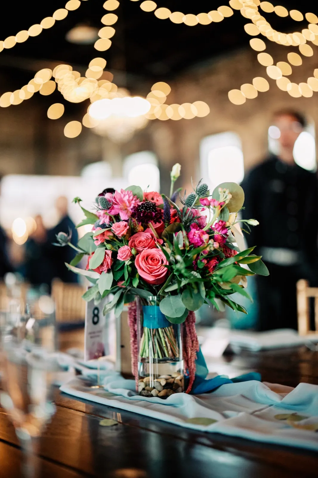A pink-and-magenta floral centerpiece stands on Maggie and Craig's wooden reception table beneath glowing string lights — Tim Larsen Photography, Brainerd Lakes MN