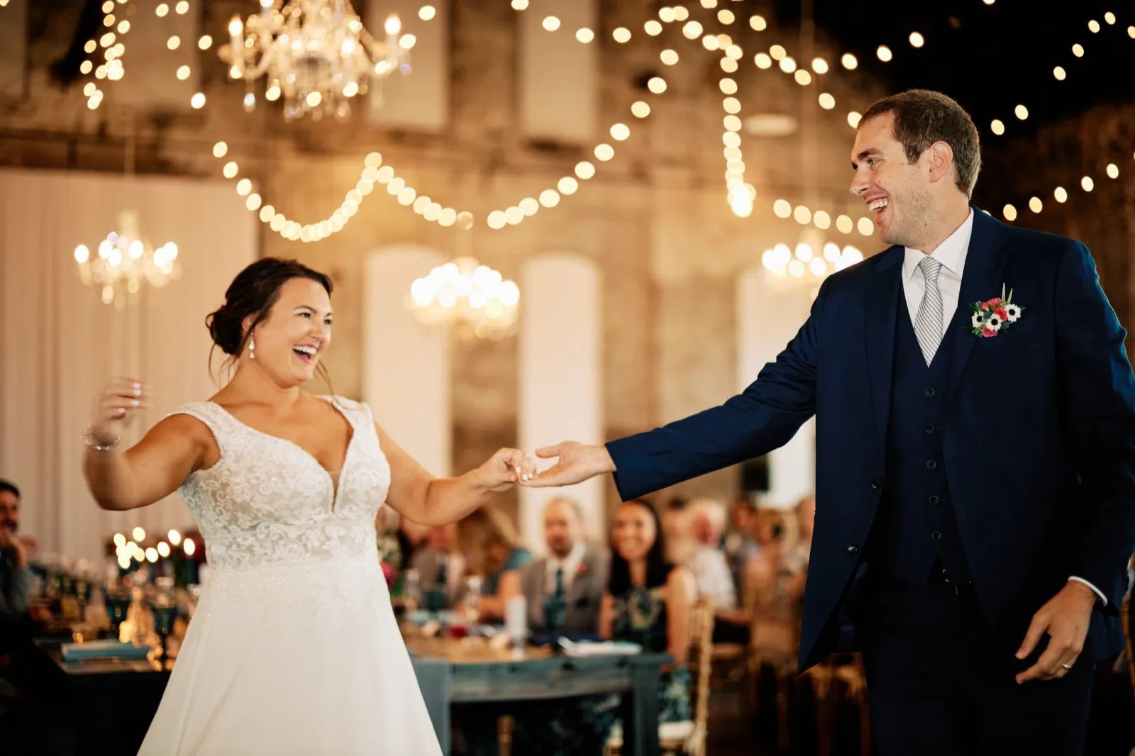 Maggie and Craig laugh with their arms outstretched during their first dance beneath chandeliers and bistro lights — Tim Larsen Photography, Brainerd Lakes MN