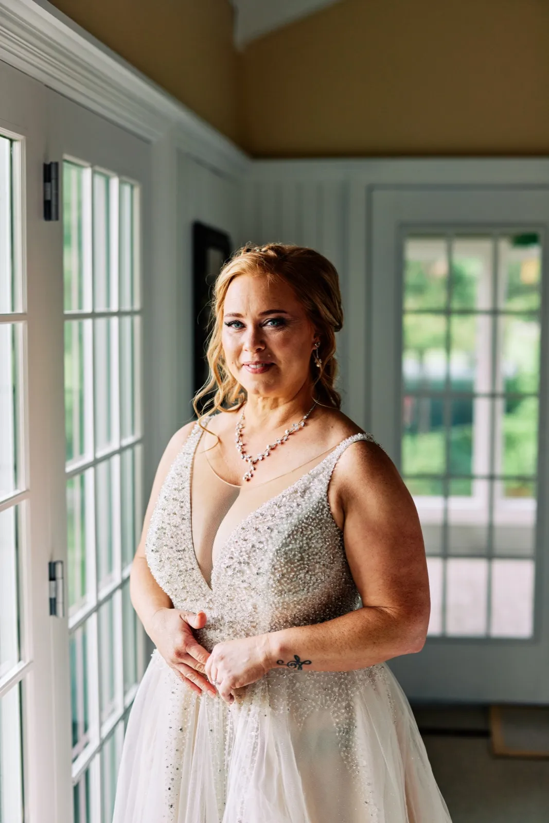 The bride stands between two french-door windows in her beaded gown, hands clasped in front of her, lit by soft afternoon window light. — Tim Larsen Photography, Brainerd Lakes MN