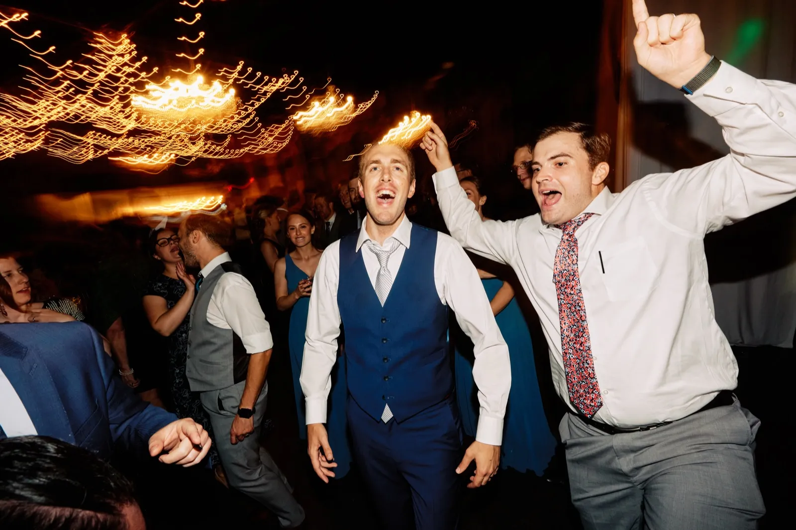 Craig celebrates on the crowded dance floor in his blue waistcoat as a guest raises an arm beside him and string lights streak overhead — Tim Larsen Photography, Brainerd Lakes MN