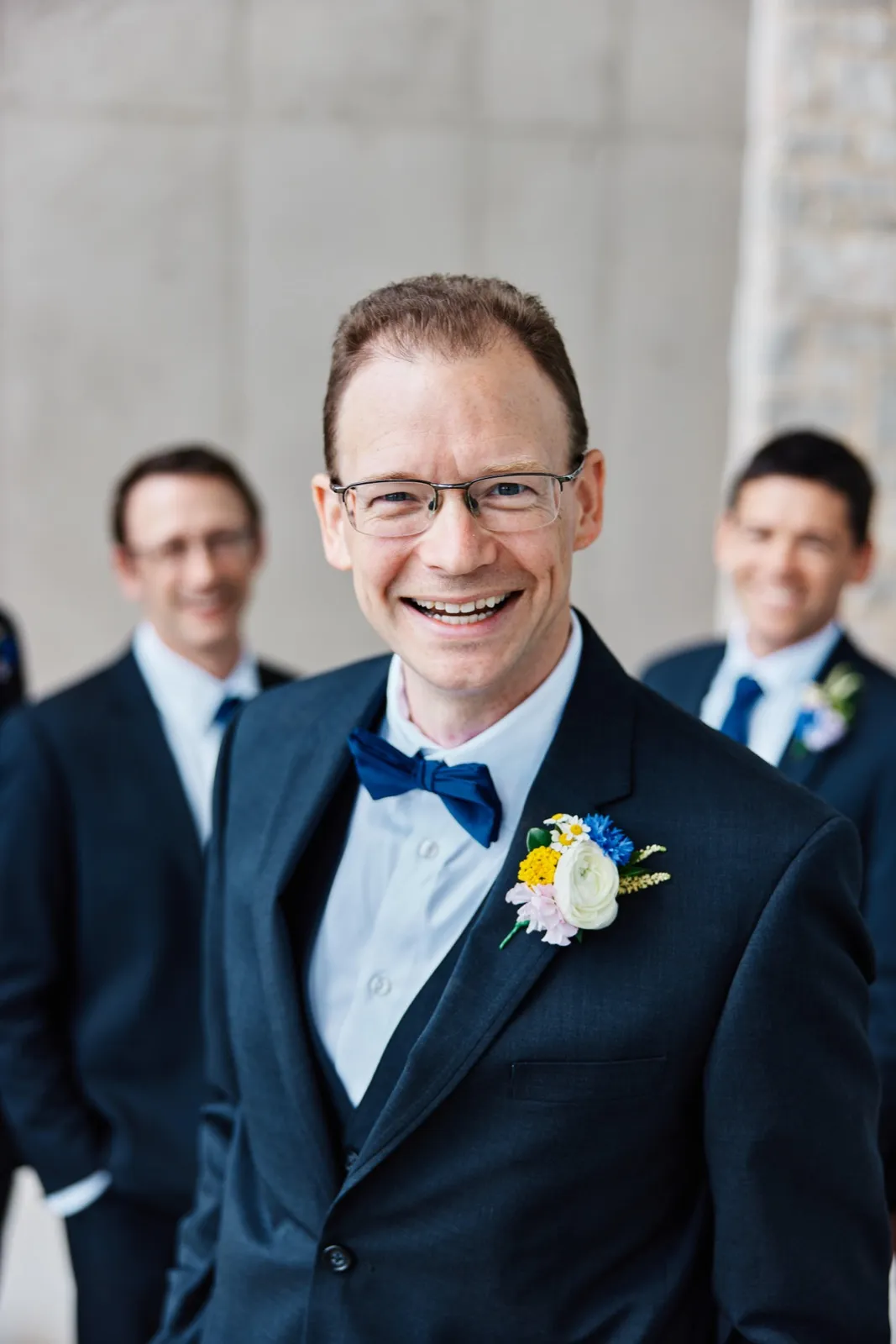 The groom smiles broadly at the camera in a navy suit and bow tie with a colorful boutonniere, two groomsmen softly out of focus behind him. — Tim Larsen Photography, Minnesota