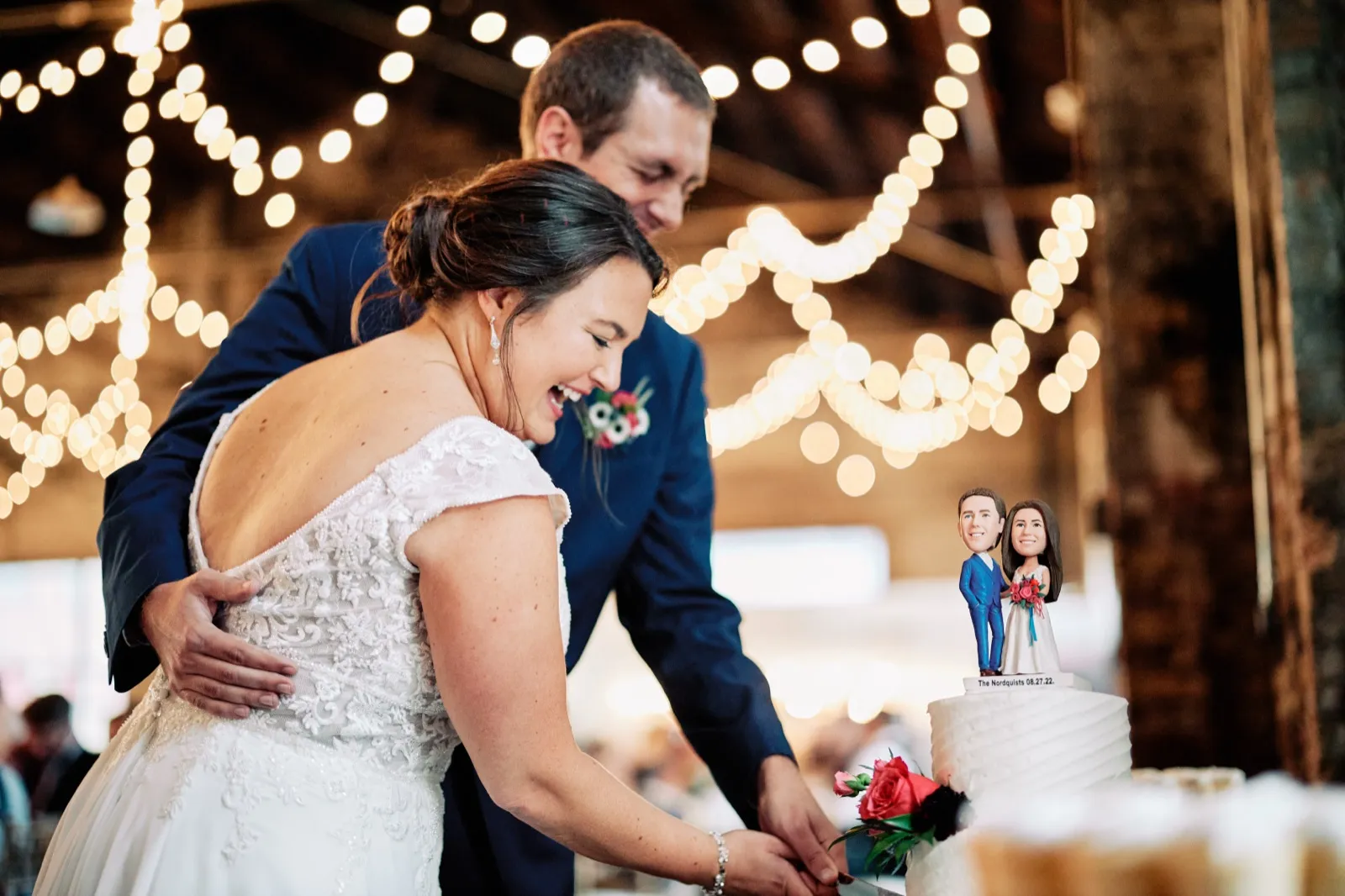 Maggie laughs as she and Craig cut their white wedding cake beneath string lights, personalized bobblehead figures on top — Tim Larsen Photography, Brainerd Lakes MN