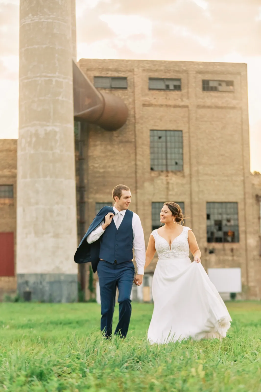 Maggie gathers her skirt and looks toward Craig as they walk across the lawn, his jacket over his shoulder and the brick powerhouse behind them — Tim Larsen Photography, Brainerd Lakes MN