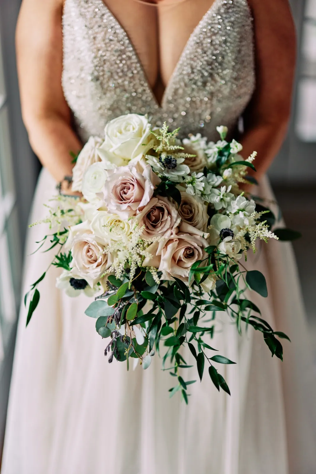 The bride holds a cascading bouquet of cream roses, blush roses, anemones, and trailing greenery against her beaded plunging-neckline gown. — Tim Larsen Photography, Brainerd Lakes MN