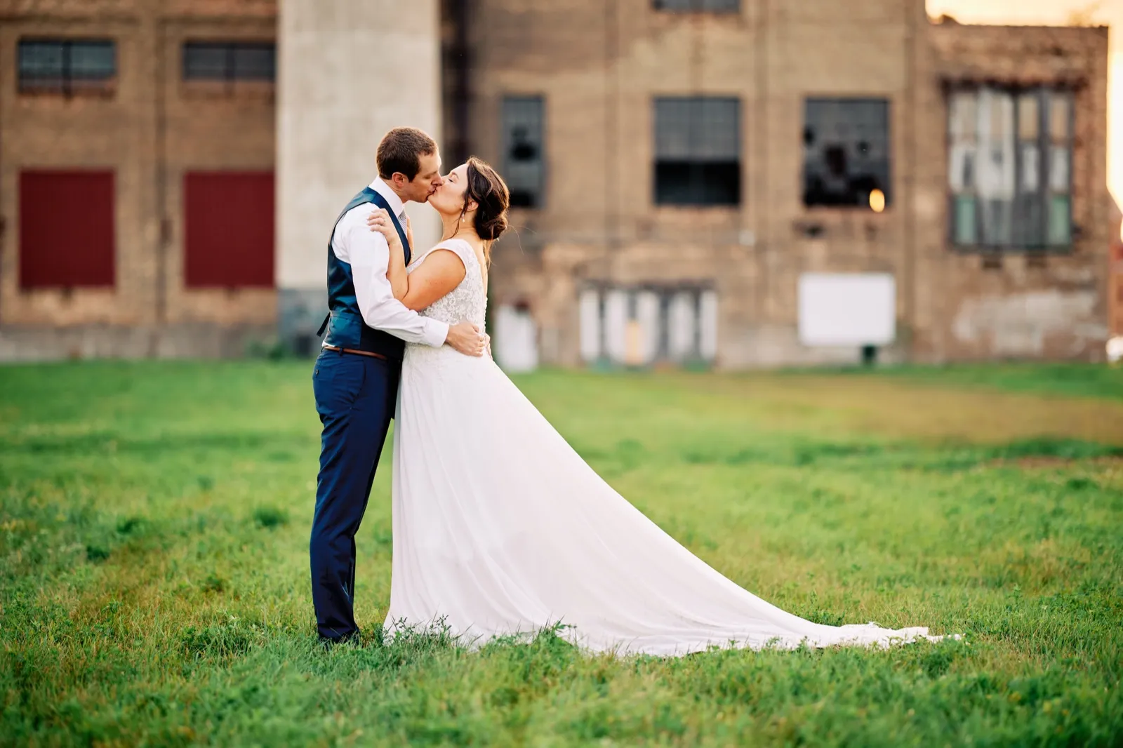 Maggie and Craig kiss on the lawn in warm evening light, her train spread across the grass in front of the brick powerhouse — Tim Larsen Photography, Brainerd Lakes MN