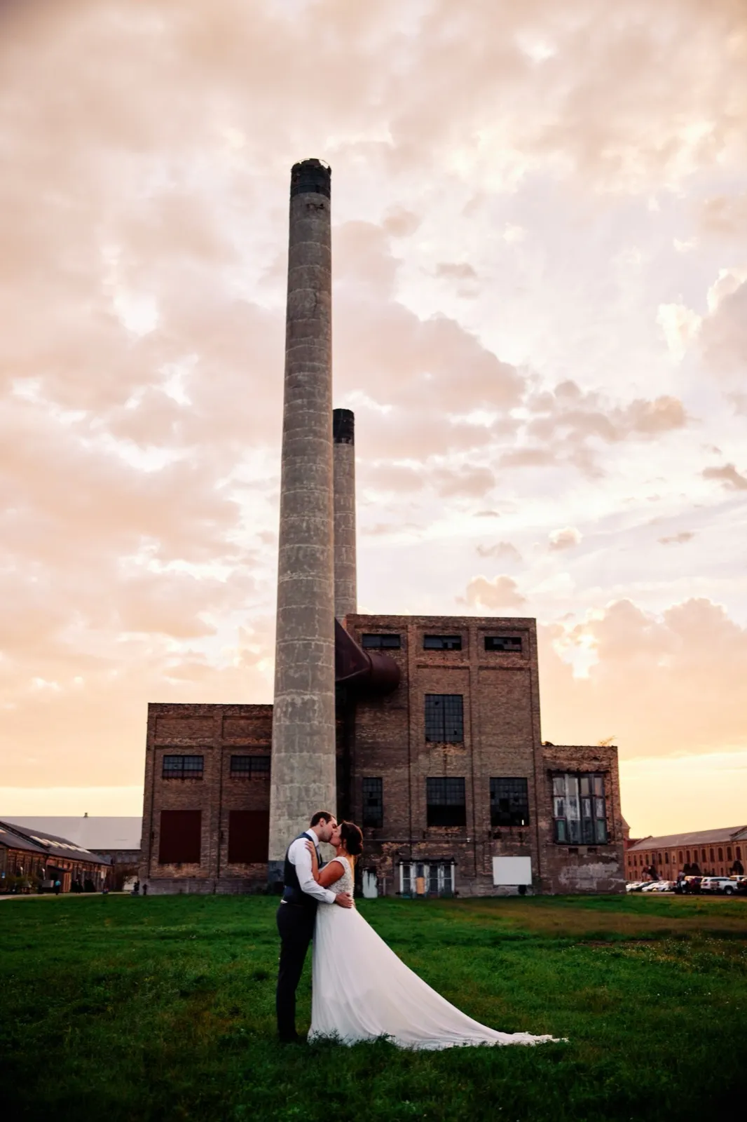 Maggie and Craig kiss on a grass lawn dwarfed by the towering brick smokestack and historic powerhouse of Northern Pacific Center beneath a streaked dusk sky — Tim Larsen Photography, Brainerd Lakes MN