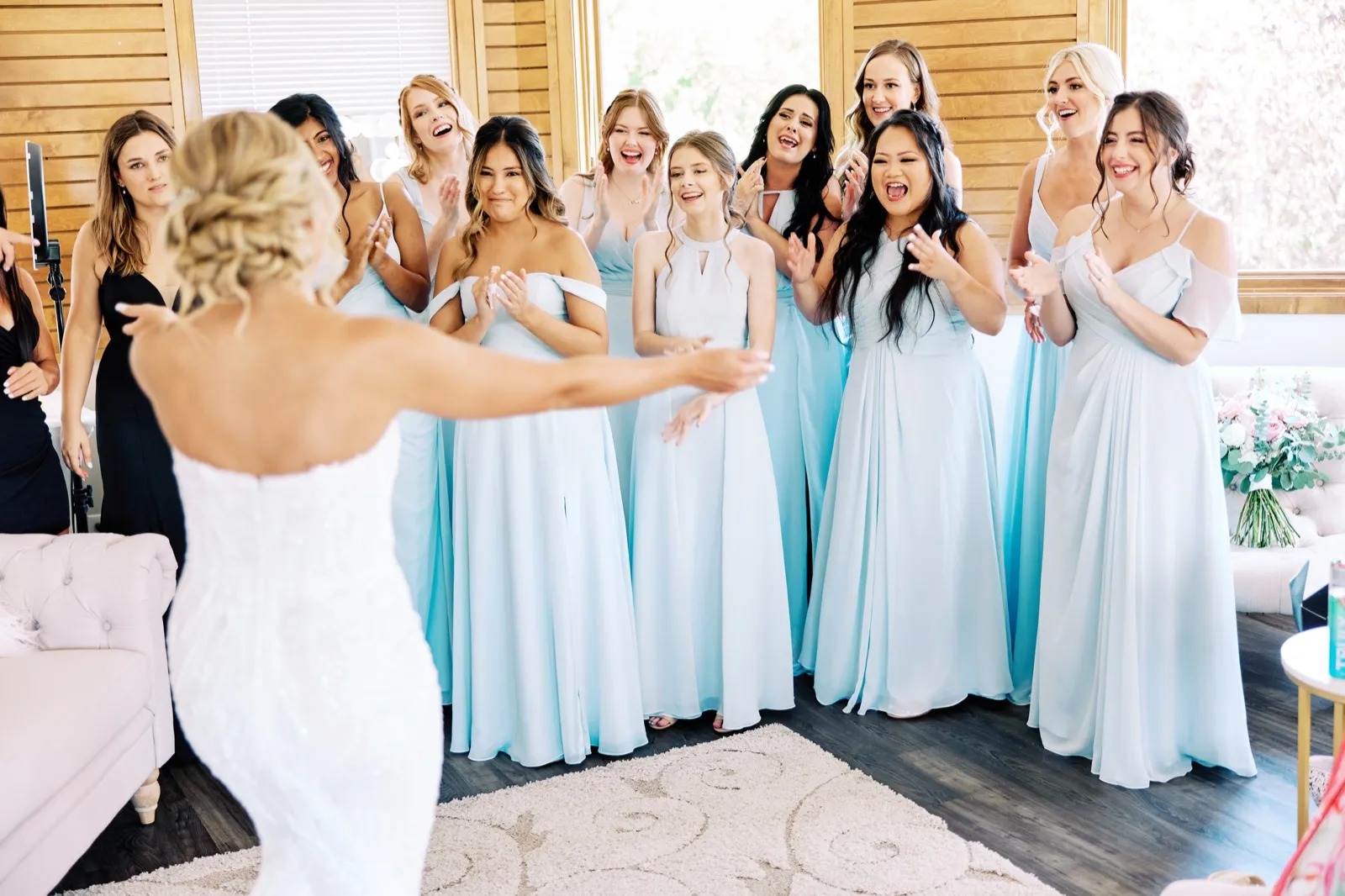 The bride stands with arms outstretched, her back to the camera, as her bridesmaids in pale blue gowns clap and react with open-mouthed delight in a sunlit bridal suite. — Tim Larsen Photography, Twin Cities MN