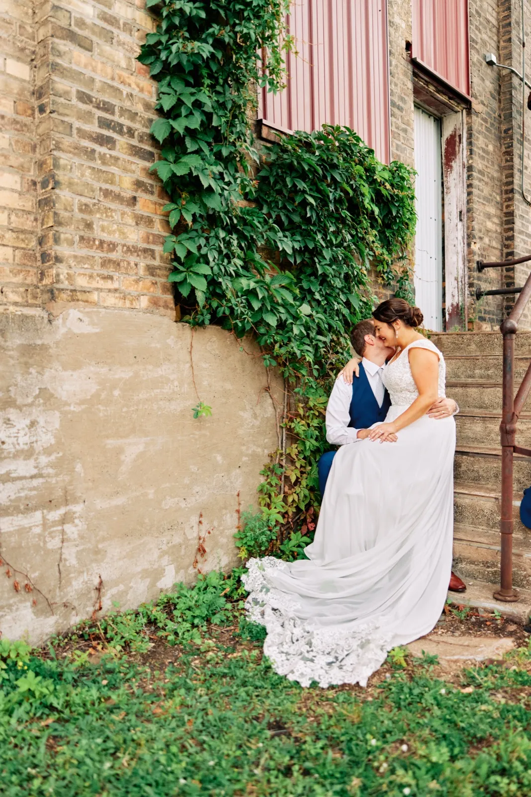 Maggie and Craig kiss beside an ivy-covered brick wall, her lace train trailing across the grass — Tim Larsen Photography, Brainerd Lakes MN