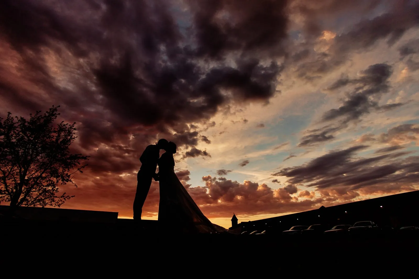 Maggie and Craig kiss in silhouette against sweeping orange and purple dusk clouds, the low rail-yard rooflines on the horizon behind them — Tim Larsen Photography, Brainerd Lakes MN
