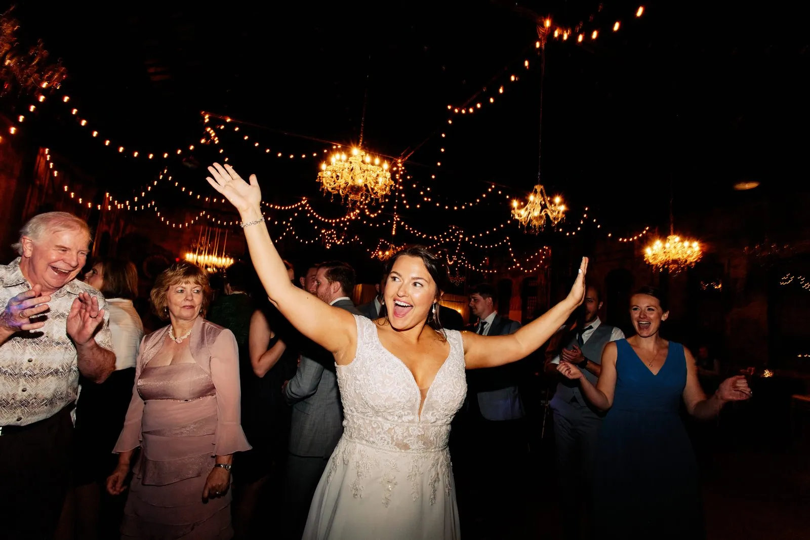 Maggie throws both arms overhead on the dance floor as guests dance around her beneath chandeliers and string lights — Tim Larsen Photography, Brainerd Lakes MN