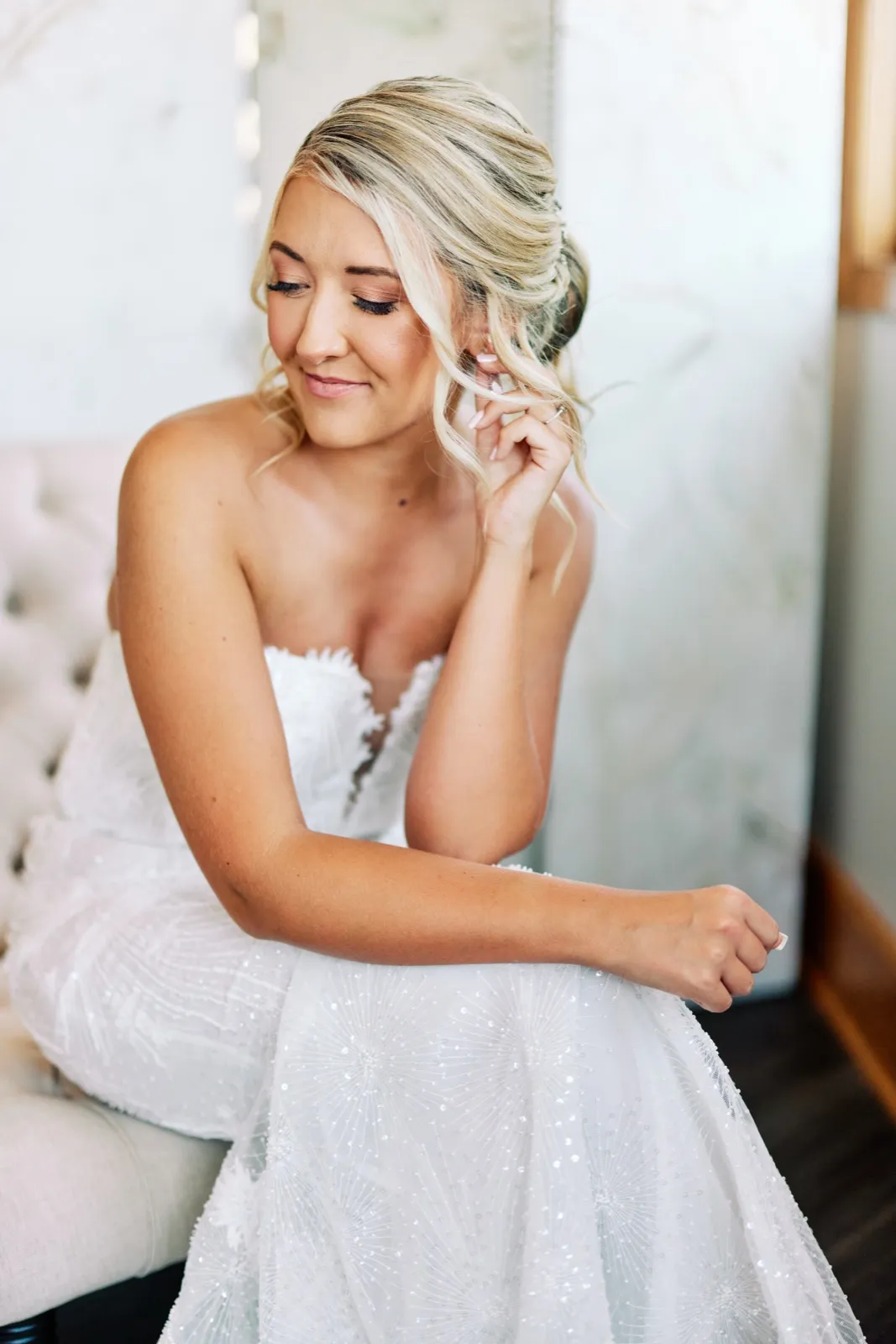 The bride sits in soft window light wearing a strapless beaded gown, gazing downward with her hand near her face. — Tim Larsen Photography, Twin Cities MN