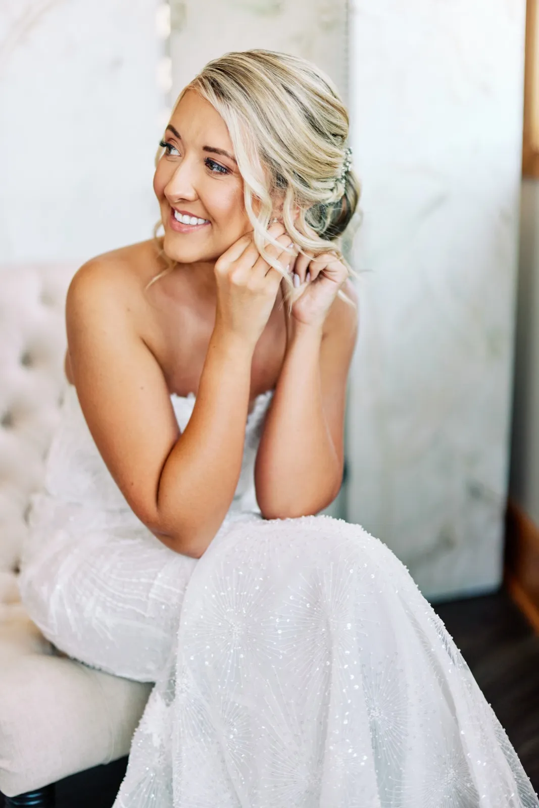 The bride sits on a tufted bench in soft window light, smiling off-camera while fastening an earring, her beaded gown cascading across her lap. — Tim Larsen Photography, Twin Cities MN